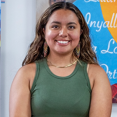 A portrait photo of a young Native American woman looking at the camera and smiling. She has long, dark hair with lighter highlights and wears a dark green tank top. 