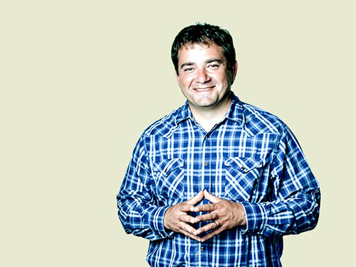 Smiling man in blue plaid shirt, fingertips together, against a buff background