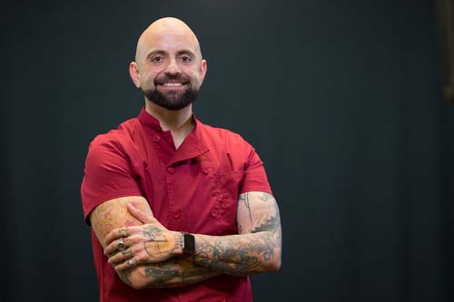 Smiling man in red shirt, heavily tattooed arms, against black background