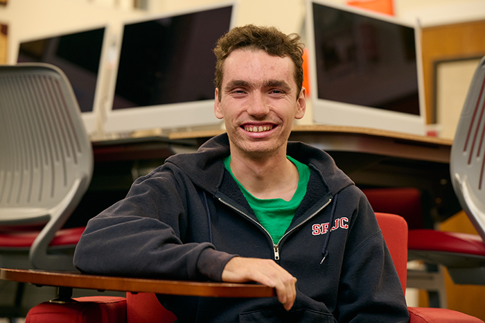 Noah McSweeney, an autistic SRJC student, smiles at the camera while sitting in the Oak Leaf Newsroom. He wears a navy blue hoodie with SRJC's logo in red and white. 
