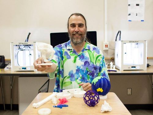 Man seated at a table covered in numerous 3D printed objects