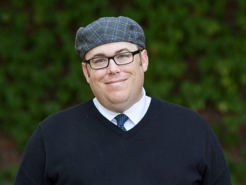 Head and shoulder portrait of a bespectacled man in front of a wall of ivy