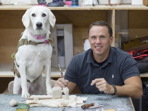 Smiling man seated at a table with art supplies, an assistance dog standing next to him