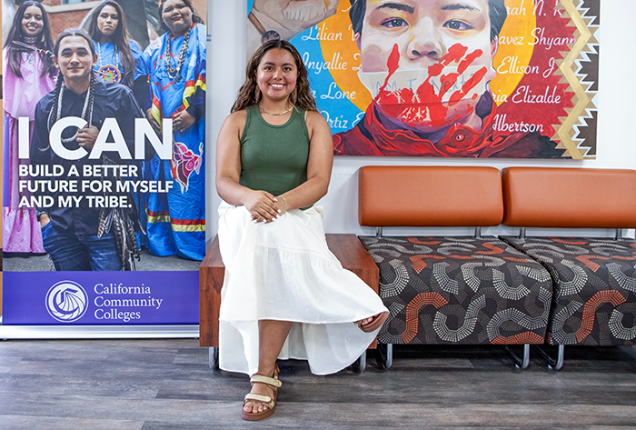 A photo of a young women seated in the Native American Center at Santa Rosa Junior College. She is wearing a green tank top and a white summer skirt. On her left and right are posters and a mural about Native American students and culture